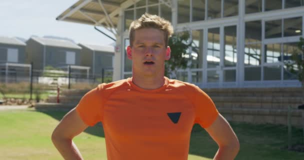 スポーツ場でオレンジのTシャツを着た白人男性ランナーの肖像画は カメラと笑顔を探して 訓練の後に立って休んでいます 陸上競技場での陸上競技トレーニング スローモーション — ストック動画