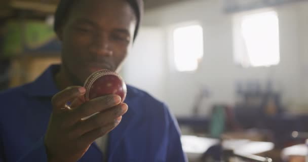 Vue de face gros plan d'un Afro-Américain travaillant dans l'atelier d'une usine fabriquant des balles de cricket, tenant et inspectant soigneusement une balle de cricket finie au soleil, au ralenti 