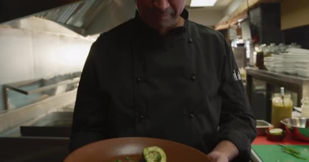 一个白人男性厨师在繁忙的餐厅厨房里工作的画像 在镜头前呈现一盘食物 面带微笑 在商业厨房工作的忙碌的厨师 — 图库视频影像
