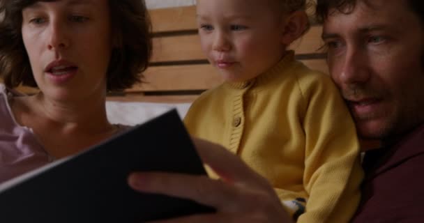 家で家族の時間を楽しんでいる白人のカップルのフロントビューを閉じる 彼らの赤ちゃんとベッドに横たわって 本を読んで スローモーション — ストック動画