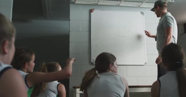 一名白人男子曲棍球教练站在旁边 与一队白人少年女子曲棍球队员交谈 并在冰球比赛前在更衣室的白板上画了一个比赛计划 一个女孩举手提问 — 图库视频影像