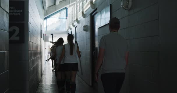 コーカシアの男子ホッケーコーチと白人の女子ホッケー選手のチームの背面ビューチームのユニフォーム変更室を残し ホッケーゲーム スローモーションの前に廊下を歩く — ストック動画