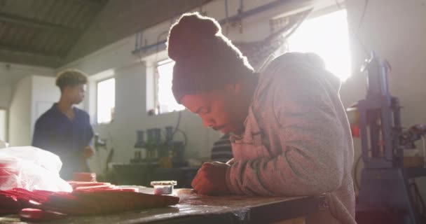 一位非洲裔美国人头戴一顶帽子 坐在工厂的工作台上 用裁剪成的红色皮革形状制作板球球 背景是同事们在生产线的其他部分工作 — 图库视频影像