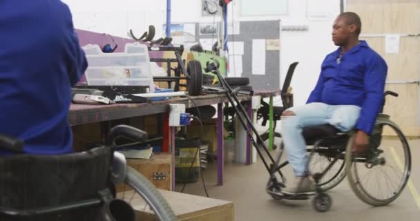 两名非洲裔美国男性工人在一家制造轮椅的工厂 在组装部分产品的工作台上的车间的侧视图 其中一名工人有残疾 并使用轮椅在工厂周围走动 — 图库视频影像