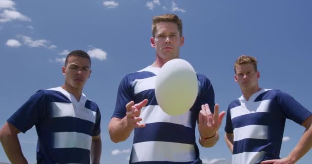 一个白人男性橄榄球手拿着球 一个白人和一个混血的男性球员站在他身后 他们都穿着队服 在赛前的橄榄球球场上 动作缓慢 — 图库视频影像