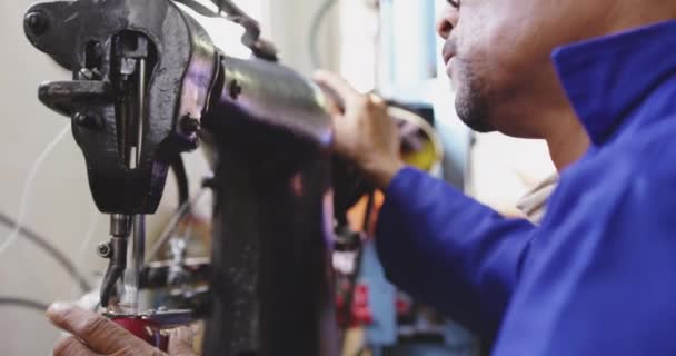 工場で働いているアフリカ系アメリカ人の男性の側のビューを閉じるクリケットボールを作るガラスを身に着けている ボールの革カバーを縫う産業用ミシンを動作します — ストック動画