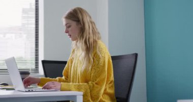 Side view of a Caucasian woman working in a creative office, working on laptop computer and drinking take away coffee