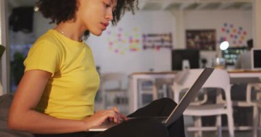 Side view of mixed race woman with curly hair and yellow t shirt working in a creative office, using laptop computer, slow motion