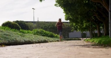 Spor kıyafetleri giymiş genç beyaz bir kadının parkta spor yaparken ağır çekimde koşması ön planda.
