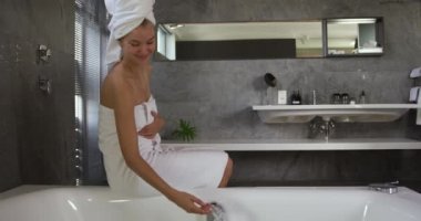 Side view of a Caucasian woman enjoying quality time Caucasian woman brushing teeth in a hotel, sitting on the side of a bathtub in bathroom and checking water,, slow motion