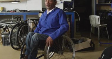 Fabrikadaki bir atölyede çalışan, tekerlekli sandalye yapan, kameraya bakıp gülümseyen ve tekerlekli sandalyede oturan Afrikalı Amerikalı bir erkek işçinin portresi.