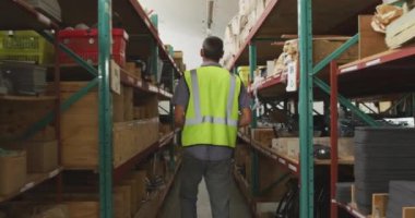 Tekerlekli sandalye üreten, yürüyen ve raflardaki parçaları inceleyen, bir tablet bilgisayar kullanan, bir depodaki beyaz erkek işçinin arka görüntüsü.