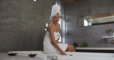 Front view of a Caucasian woman enjoying quality time in a hotel, sitting on the side of a bathtub in bathroom, slow motion