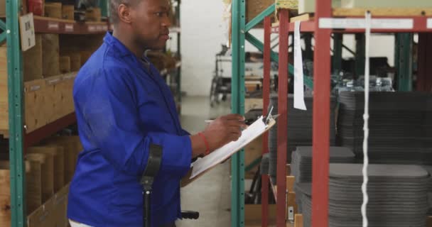 在一家制造轮椅的工厂的仓库里 一个非洲裔美国男性员工的侧视图 他在剪贴板上写字 看着相机 微笑着 站在拐杖上 — 图库视频影像