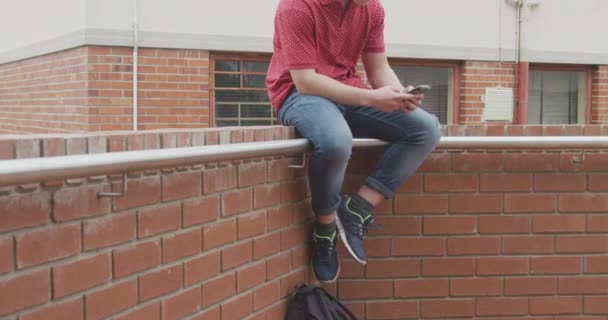 在阳光灿烂的日子里 一个白人小学生坐在校园的墙上 慢吞吞地听着耳机里的音乐 — 图库视频影像