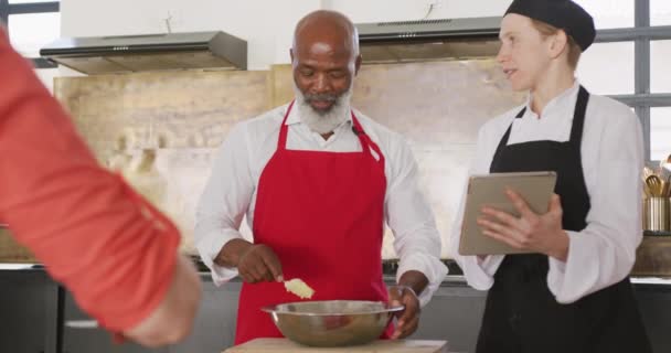 レストランのキッチンで料理教室をしているアフリカ系アメリカ人の先輩の前で 白人のシェフを着てタブレットを使っている白人女性シェフの指示に耳を傾け スローモーションで — ストック動画