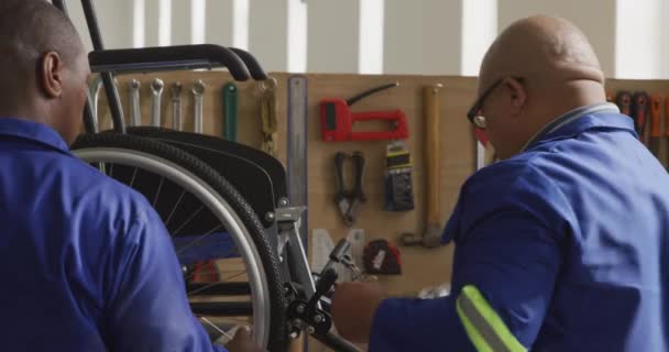 一位非洲裔美国人和一位混血男工在一家制造轮椅的工厂的车间里 站在工作台上忙着组装轮椅零件 面前挂着工具的侧视图 — 图库视频影像