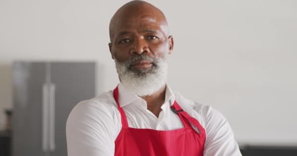 赤いエプロンを着て腕を組んで料理教室に立ちカメラを向け笑顔でゆっくりと動き回るアフリカ系アメリカ人の先輩の肖像画 — ストック動画