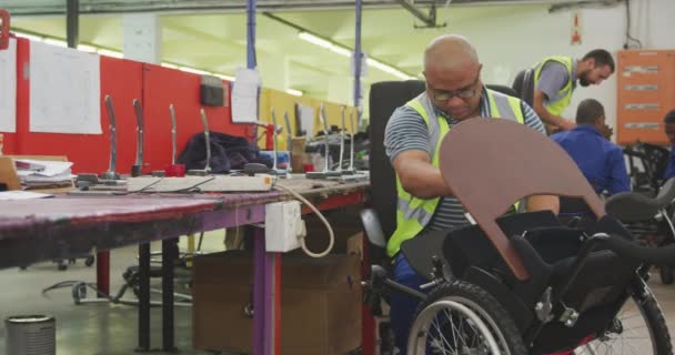 車椅子を作る工場でのワークショップでの混合レースの男性労働者のフロントビュー 車椅子の一部を組み立て作業台に座ってトレイに置き 車輪をチェック — ストック動画