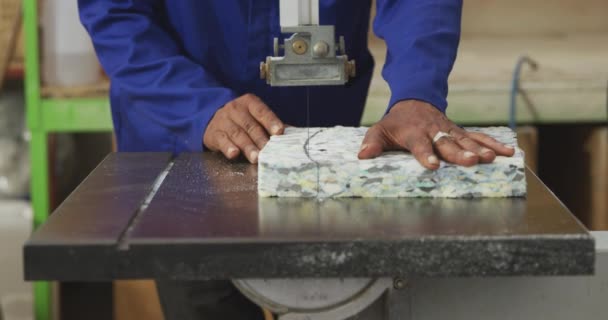 在一家制造轮椅的工厂的车间里 一位非洲裔美国男性工人站在工作台上 用带锯割破一块泡沫 他的镜头正中 — 图库视频影像