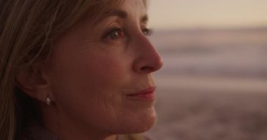 Son sınıf beyaz bir kadının gün batımında kumsalda oturup yavaşça denize gülümseyen ve hayran olan doğanın tadını çıkarırken yan görüntüsü.