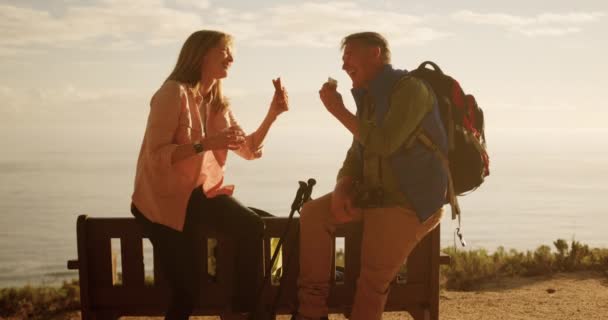一对年长的白种人夫妇在一起享受大自然的时光 一边在悬崖上坐着 一边用北欧手杖走路 一边笑 一边慢吞吞地吃三明治的侧影 — 图库视频影像