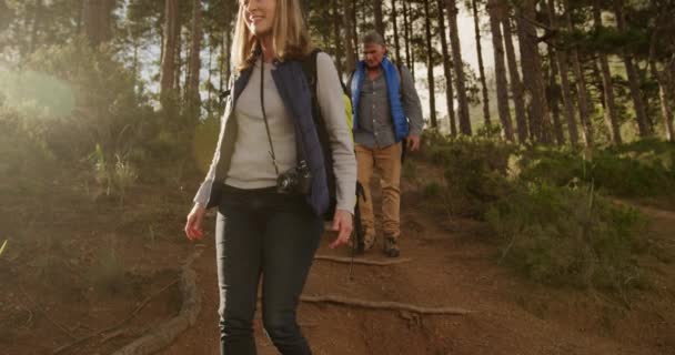 自然の中で一緒に時間を楽しむ先輩白人夫婦のフロントビュー バックパックやカメラとハイキング中に森の中を歩く スローモーションで笑顔 — ストック動画