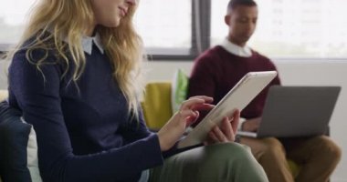 Yaratıcı bir ofiste çalışan, lobide oturan, arka planda dizüstü bilgisayarda çalışan melez erkek arkadaşıyla tablet bilgisayar kullanan beyaz bir kadının yan görüntüsü.