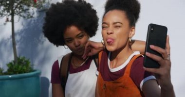 Güneşli bir günde sokakta boş vakit geçiren, akıllı telefonuyla selfie çeken iki melez kadının ön görüntüsü.