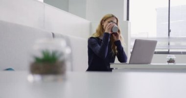 Yaratıcı bir ofiste çalışan, akıllı telefonla konuşan, dizüstü bilgisayar ekranına bakan, kahve içen beyaz bir kadının yan görüntüsü.