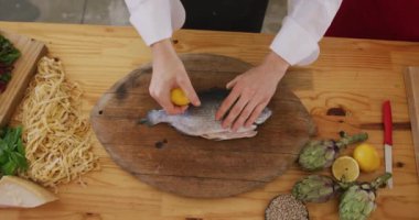 Restoran mutfağında aşçılık kursu sırasında beyaz bir kadın şefin ellerini üst açıda görmek için yüksek açılı orta açı, yavaş çekimde pişirmeye hazırlanan çiğ balığın üzerine limon suyu sıkmak.