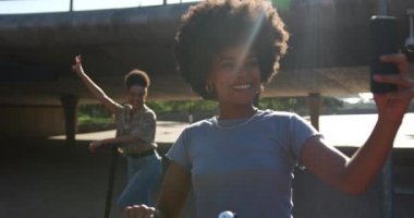 Güneşli bir günde şehir parkında eğlenen iki melez kadının ön görüntüsü. Elektrikli scooterlara binmek, fotoğraf çekmek, ağır çekim...