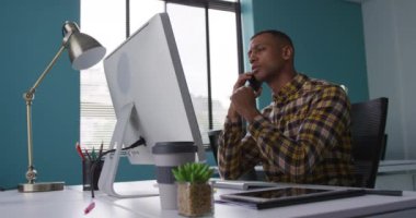 Yaratıcı bir ofiste çalışan, akıllı telefonla konuşan, bilgisayar ekranına bakan, ağır çekimde çalışan melez bir adamın yan görüntüsü.