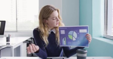 Yaratıcı bir ofiste çalışan, kameraya konuşan, grafikleri ve istatistikleri gösteren, gülümseyen mutlu, beyaz bir kadının ön görüntüsü.