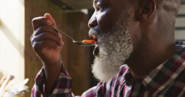 Restoran mutfağındaki aşçılık sınıfında beyaz sakallı mutlu bir Afrikalı Amerikalı adamın yan görüntüsü. Yavaş çekimde kaşıktan sıcak bir yemeğin tadını çıkarıyor.