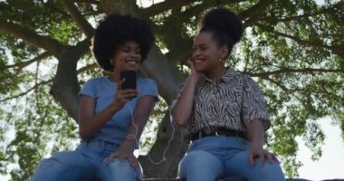 Güneşli bir günde şehir parkında boş vakit geçiren iki melez kadının düşük açılı görüşü, akıllı telefon kullanımı, kulaklıkları paylaşması ve müzik dinlemesi, yavaş çekim.