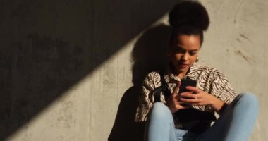 Güneşli bir günde şehir parkında boş vakit geçiren, akıllı telefon kullanan, yavaş çekim yapan melez bir kadının ön görüntüsü.