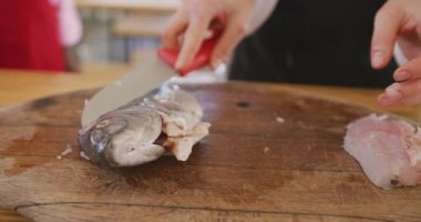Restoran mutfağındaki aşçılık sınıfında beyaz şefler ve siyah önlükler giyen beyaz bir kadın şefin orta kesiminde yetişkin öğrencilere ağır çekimde çiğ balığı nasıl keseceklerini gösteriyor.