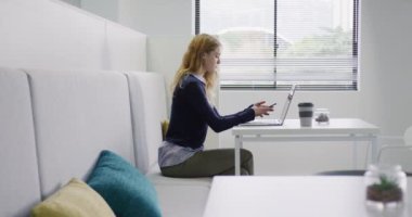 Yaratıcı bir ofiste çalışan, akıllı telefonla konuşan ve dizüstü bilgisayar ekranına bakan beyaz bir kadının yan görüntüsü.