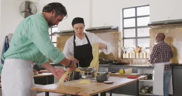 在餐馆厨房的烹饪课上 一位高个子白人男子和一位白人女厨师的侧视图 厨师慢动作地解释并帮助这名男子通过面食机滚动面团 — 图库视频影像