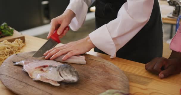 レストランのキッチンで調理教室中に白人女性シェフのサイドビューの真ん中のセクションでは 木製のまな板に鋭いナイフで新鮮な魚を記入し スローモーションで — ストック動画