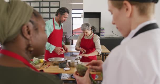 料理教室で一緒に料理を作る大人の白人男性と女性の正面図 アフリカ系アメリカ人女性が前景に白人女性シェフと話しながら 背景に焦点を当て スローモーションで — ストック動画