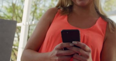 Genç, beyaz bir kadının yaz tatilinde balkondaki bir masada oturup, yavaş çekimde akıllı telefon kullanmasının ön görüntüsü..