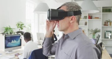 Kafkasyalı bir işadamının modern bir ofiste çalışmasının yan görüntüsü. Sanal gerçeklik kulaklığı takıyor ve meslektaşları arka planda ağır çekimde çalışıyor.