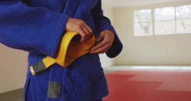 Beyaz kadın judoka 'nın orta kesiminde spor salonunun paspasları üzerinde duruyor, mavi judoginin sarı kuşağını yavaş çekimde bağlıyor.