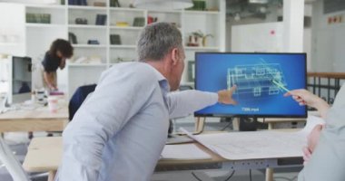 Modern bir ofiste çalışan iki beyaz mimarın yan görünüşü, bir masanın başında oturmuş, 3D mimari çizimle iş arkadaşlarıyla arka planda yavaş çekimde çalışırken beyin fırtınası yapmayı tartışıyorlar.