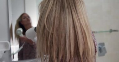 Kafkasyalı genç bir kadının, uzun, sarı saçlarını saç kurutma makinesiyle kuruturken, zarif bir otel odasında, yavaş çekimde, boş zamanlarının tadını çıkarırken görüntüsü..