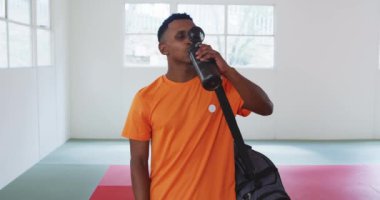 Kendinden emin, mutlu, turuncu tişört giyen, spor salonunda spor çantasıyla duran, judo eğitiminden sonra plastik şişeden su içen genç erkek judoka 'nın ön görüntüsü..