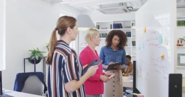 Modern bir ofiste çalışan üç çok ırklı iş kadınının yan görünüşü, beyaz tahta başında oturmuş, erkek meslektaşlarına masasında oturup bilgisayar ekranlarına bakarak beyin fırtınası yapıyor ve yazıyorlar.