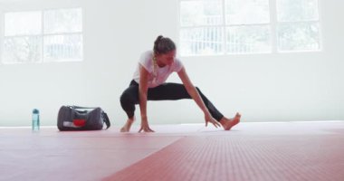Beyaz, genç bir kadın judoka 'nın yan görüntüsü, jimnastik paspaslarında ısınma, judo antrenmanından önce bacaklarını esnetme, arka planda ağır çekimde spor çantası..